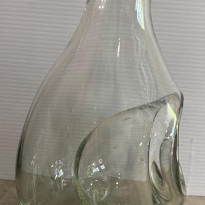 Puede incluir: Una jarra de vidrio transparente con un diseño asimétrico único. La jarra tiene una base ancha que se estrecha hacia un cuello estrecho y un borde acampanado. Una forma grande y redondeada en forma de burbuja está unida al costado.