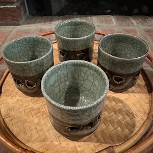May include: Four ceramic cups with a crackle glaze in shades of blue and gray, set on a round bamboo tray. The cups have a dark brown band with decorative cutouts. The cups are likely for tea or coffee.