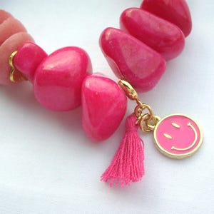 May include: A close-up of a pink beaded bracelet with a gold charm and tassel. The bracelet features large, polished pink beads and smaller, round pink beads. The gold charm is a pink smiley face.