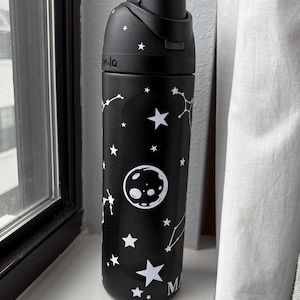 Puede incluir: Botella de agua negra con diseño de estrellas blancas, constelaciones y una luna. La botella tiene una tapa negra con apertura de botón. Diseño con temática celestial.