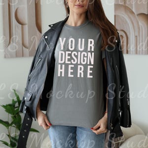 May include: A gray t-shirt with the text "YOUR DESIGN HERE" in light pink, worn with a black leather jacket and blue jeans. The image is a mockup, showcasing a design on a t-shirt.