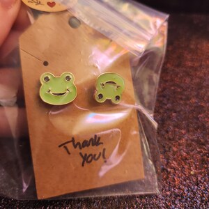 May include: A pair of green frog-shaped stud earrings. The earrings are in a clear plastic bag, attached to a brown tag that says "Thank You!" in black ink. The earrings have a black outline and a smiling face.