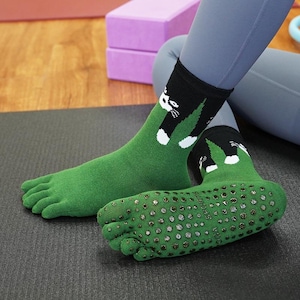 May include: Green toe socks with a black cuff featuring a white cat design. The soles have small, non-slip grips. The socks are designed for yoga or similar activities.