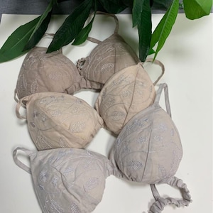 May include: A selection of beige and light grey bras, each featuring embroidered leaf designs. The bras have adjustable straps and are arranged on a white surface. Green leaves are visible in the background.