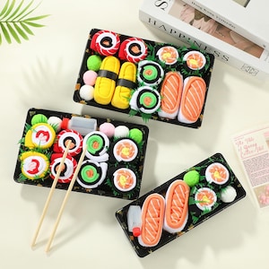 May include: Three black rectangular trays containing rolled socks designed to resemble various sushi pieces, including nigiri and maki rolls. The sets feature vibrant colours like orange, yellow, green, and white, with wooden chopsticks included.