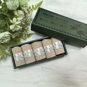 以下が含まれることがあります： ベージュ色のヘンプソックス5足セット。緑色の箱に入っています。箱の蓋には「HEMP SOCKS」の文字と大麻の葉のイラストが描かれています。各靴下は「HEMP」と書かれたラベルで包まれており、大麻の植物のデザインが特徴です。