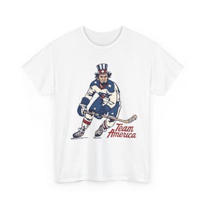 Könnte beinhalten: Weißes T-Shirt mit einer patriotischen Illustration eines Eishockeyspielers in Rot, Weiß und Blau. Das Design beinhaltet einen Zylinder und den Text "Team America" in roter Schrift. Für Sportfans.