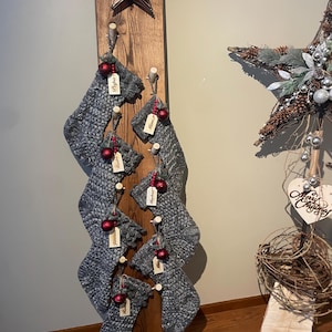 May include: A wooden Christmas stocking holder with six gray knit stockings, each adorned with a red ornament and a name tag. A decorative star is at the top, and a larger star is to the right.