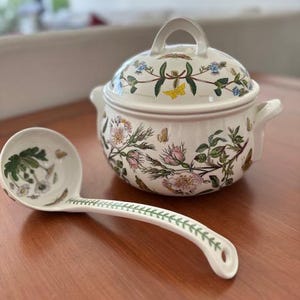 May include: A white ceramic soup tureen with a matching ladle. The tureen has a lid and handles, and both pieces are decorated with floral and butterfly patterns in pink, green, and yellow. The ladle has a long handle with a green leaf design.