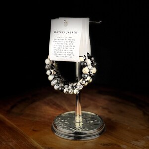 May include: A Matrix Jasper beaded bracelet displayed on a silver stand. The bracelet features black and white patterned beads. A white card with the text "MATRIX JASPER" and its properties is attached to the stand.