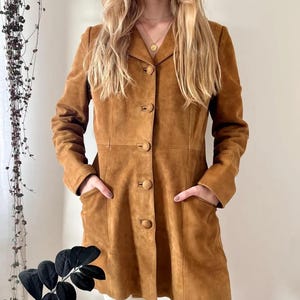 Può includere: Un cappotto in camoscio marrone chiaro con chiusura a bottoni e due tasche anteriori. Il cappotto ha un colletto classico e una vestibilità su misura. La modella indossa il cappotto con pantaloni bianchi. Il cappotto è di media lunghezza.