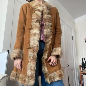 Op de afbeelding: Een lange, bruine suède jas met imitatiebont langs de kraag, manchetten en voorkant. De jas heeft knevelsluitingen en een vintage uitstraling. De bontafwerking is lichtbruin.