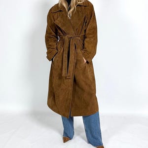May include: A long, brown suede coat with a belted waist and large lapels. The coat is knee-length and has pockets. The model is wearing blue jeans and brown pointed-toe heels. A classic outerwear garment.