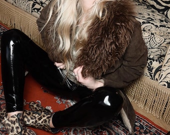 Puede incluir: Una chaqueta de ante marrón con cuello de piel se combina con leggings negros brillantes de cuero sintético. El conjunto se completa con botines con estampado de leopardo. La estética general es de inspiración vintage y atrevida.