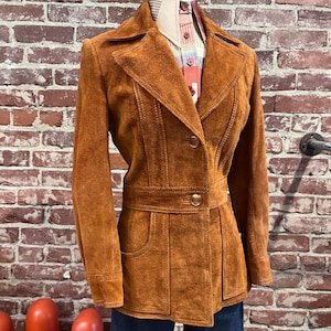 Könnte beinhalten: Vintage braune Wildlederjacke mit tailliertem Schnitt, mit Reverskragen, Knopfverschluss und Taillengürtel. Die Jacke hat zwei Vordertaschen und wird auf einer Schaufensterpuppe präsentiert. Die Jacke wird mit einem gemusterten Hemd und blauen Jeans kombiniert.
