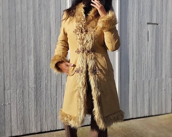 May include: A long, tan coat with faux fur trim on the collar, cuffs, and hem. The coat features decorative floral embroidery and ornate closures down the front. The coat is made of a soft, suede-like material, offering a vintage aesthetic.