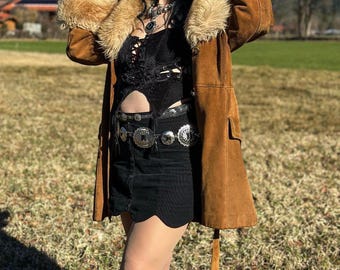 Puede incluir: Una chaqueta de gamuza marrón con cuello de piel sintética y un top de encaje negro debajo. La chaqueta tiene un cinturón con detalles de concho plateados.