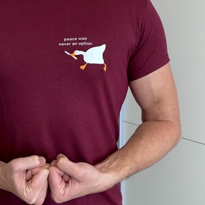 Può includere: Una maglietta color granata con un'oca bianca che tiene un coltello e il testo "peace was never an option".