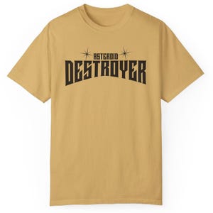 May include: Mustard-colored t-shirt with the text "ASTEROID DESTROYER" in bold, black, stylized font. The text is arched, with starburst designs above. The shirt has short sleeves and a crew neck.