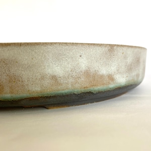 May include: A handcrafted ceramic bowl with a wide, shallow design. The bowl has a speckled, off-white glaze with brown and green accents near the rim. The base is a dark brown colour.