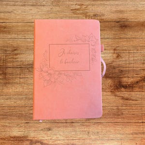 Peut inclure: Un carnet de couleur corail avec une bordure rectangulaire et des motifs floraux au trait sur la couverture. Le texte "Je choisis le bonheur" est imprimé au centre. Le carnet dispose d'une bande élastique assortie sur le côté droit.