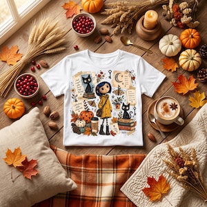 Könnte beinhalten: Ein weißes kurzärmeliges T-Shirt mit einer grafischen Illustration einer Figur im gelben Regenmantel, schwarzen Katzen, Kürbissen, Herbstblättern und dem Text Just a girl who loves cozy things, Collecting little moments und Dear girl, more cozy days.