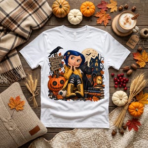 Könnte beinhalten: Ein weißes T-Shirt mit einer grafischen Illustration einer blauhaarigen Figur in einem gelben Regenmantel, die neben einer schwarzen Katze mit Knopfaugen sitzt. Das Design zeigt ein Holzschild mit der Aufschrift Somewhere Other World, einen Kürbis und ein Haus.