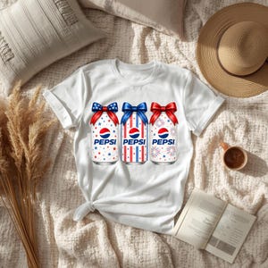 Könnte beinhalten: Weißes T-Shirt mit drei Pepsi-Dosen mit patriotischen Designs. Jede Dose hat ein rot-weiß-blaues Thema, verziert mit Schleifen und dem Pepsi-Logo. Die Dosen sind mit Sternen, Streifen und Feuerwerk verziert.