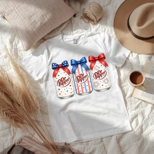 Könnte beinhalten: Weißes T-Shirt mit drei Dr. Pepper-Dosen im patriotischen Design. Jede Dose hat eine rot-weiß-blaue Schleife mit Stern-, Streifen- und Feuerwerksgrafiken. Das Dr. Pepper-Logo ist auf jeder Dose sichtbar.