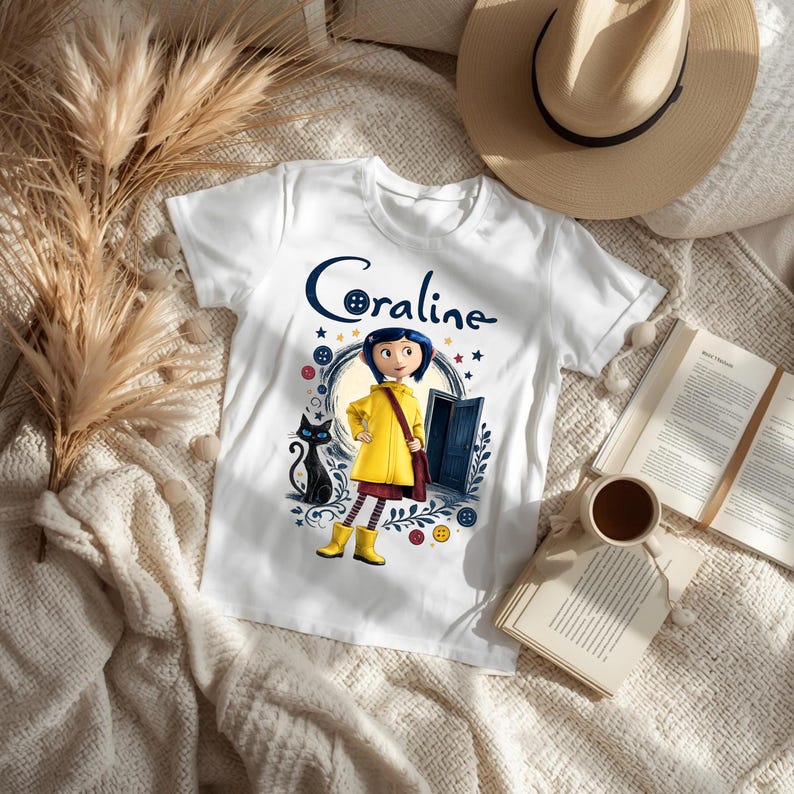 K&ouml;nnte beinhalten: Ein wei&szlig;es kurz&auml;rmeliges T-Shirt mit einem Grafikdruck der Figur Coraline in gelbem Regenmantel und Stiefeln, begleitet von einer schwarzen Katze, einer blauen T&uuml;r und dem Schriftzug Coraline, bei dem ein Knopf den Buchstaben o ersetzt.