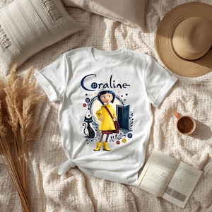 K&ouml;nnte beinhalten: Ein wei&szlig;es kurz&auml;rmeliges T-Shirt mit einem Grafikdruck der Figur Coraline in einem gelben Regenmantel und Stiefeln, begleitet von einer schwarzen Katze, einer blauen T&uuml;r und bunten Kn&ouml;pfen, mit dem Text Coraline &uuml;ber der Figur gedruckt.