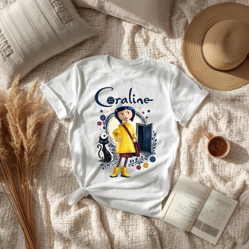 K&ouml;nnte beinhalten: Ein wei&szlig;es kurz&auml;rmeliges T-Shirt mit einem Grafikdruck der Figur Coraline in einem gelben Regenmantel und Stiefeln, begleitet von einer schwarzen Katze, einer blauen T&uuml;r und bunten Kn&ouml;pfen, mit dem Text Coraline &uuml;ber der Figur gedruckt.