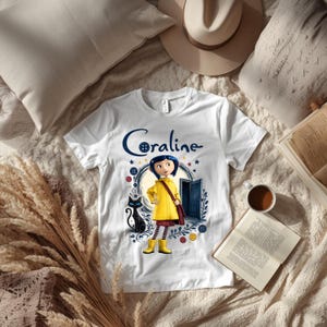 K&ouml;nnte beinhalten: Ein wei&szlig;es Kurzarm-T-Shirt mit einem Grafikdruck der Figur Coraline, die einen gelben Regenmantel und Stiefel tr&auml;gt und neben einer schwarzen Katze steht. Das Design enth&auml;lt eine blaue T&uuml;r, bunte Kn&ouml;pfe und den Schriftzug Coraline in Blau.