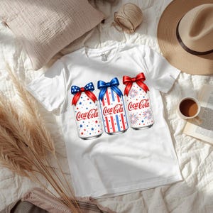 Puede incluir: Camiseta blanca con un diseño de tres latas de Coca-Cola decoradas con motivos patrióticos. Cada lata tiene un lazo y está decorada con estrellas, rayas y fuegos artificiales rojos, blancos y azules. El logotipo de Coca-Cola es visible.