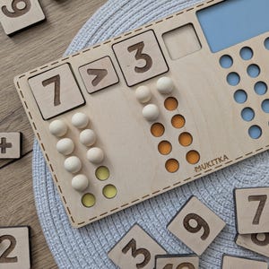 Pu&ograve; includere: Giocattolo educativo in legno con un tabellone numerico, gettoni e un cartello che dice "MUKITKA". Il tabellone ha i numeri 3, 7 e un simbolo maggiore di. Ci sono anche gettoni nei colori giallo e arancione.