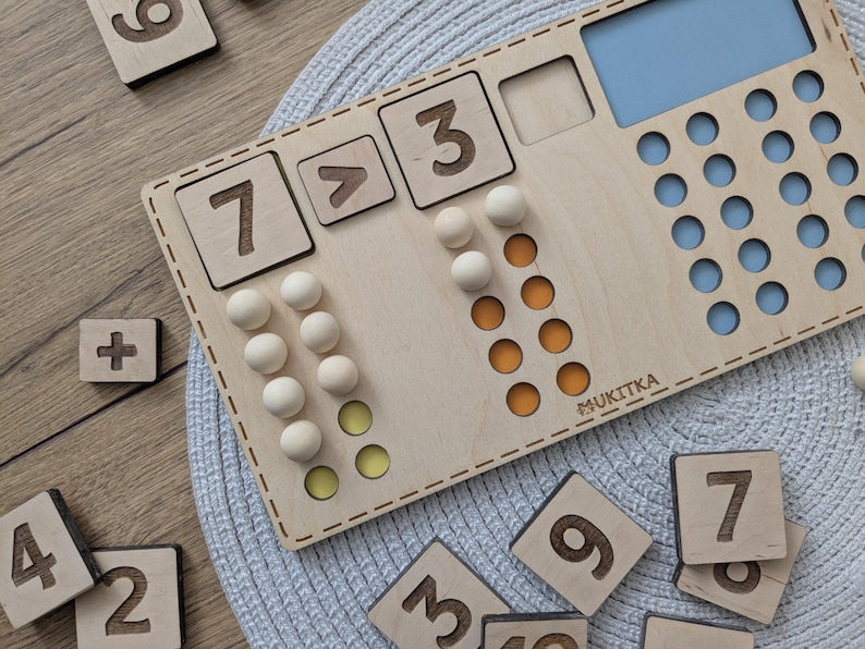 Pu&ograve; includere: Giocattolo educativo in legno con un tabellone numerico, gettoni e un cartello che dice "MUKITKA". Il tabellone ha i numeri 3, 7 e un simbolo maggiore di. Ci sono anche gettoni nei colori giallo e arancione.