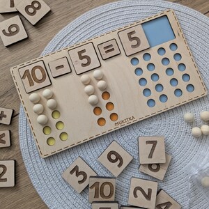 Pu&ograve; includere: Giocattolo educativo in legno con un tabellone numerico e dei contatori. Il tabellone mostra l'equazione 10 - 5 = 5. Ci sono 10 contatori in legno in totale, 5 dei quali sono gialli e 5 dei quali sono arancioni. Il tabellone &egrave; fatto di legno chiaro e ha uno sfondo blu e bianco. Il tabellone &egrave; etichettato "MUKITKA".