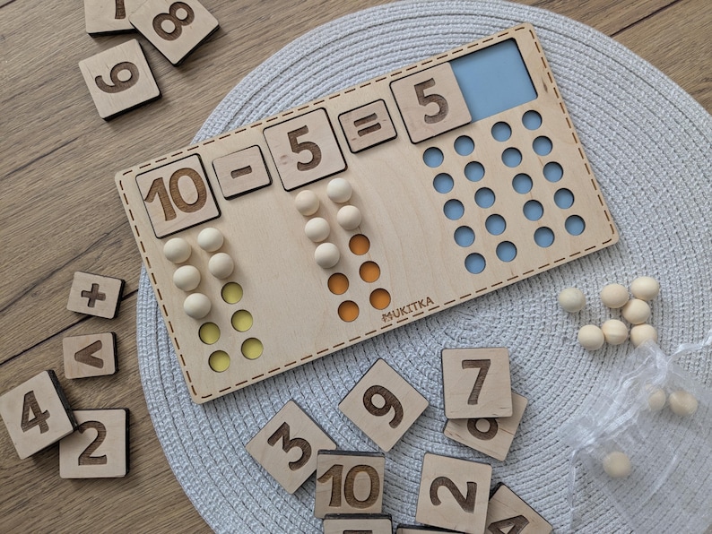Pu&ograve; includere: Giocattolo educativo in legno con un tabellone numerico e dei contatori. Il tabellone mostra l'equazione 10 - 5 = 5. Ci sono 10 contatori in legno in totale, 5 dei quali sono gialli e 5 dei quali sono arancioni. Il tabellone &egrave; fatto di legno chiaro e ha uno sfondo blu e bianco. Il tabellone &egrave; etichettato "MUKITKA".