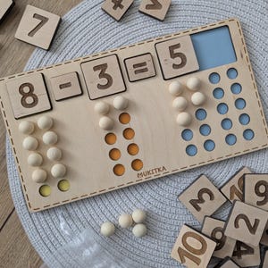 以下が含まれることがあります： 数字のボードとビーズが付いた木製教育玩具。ボードには8 - 3 = 5という式が示されています。最初の列には8個のビーズ、2番目の列には3個のビーズ、3番目の列には5個のビーズがあります。また、ビーズと数字のタイルも含まれています。