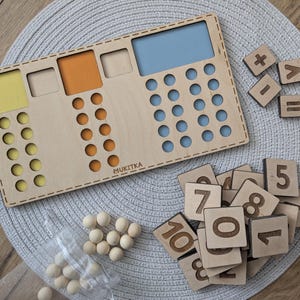 Pu&ograve; includere: Un tabellone di conteggio in legno con sezioni colorate e un'area blu per contare. Il tabellone include un set di tessere numeriche in legno, segni pi&ugrave;, meno, uguale e maggiore di. Ci sono anche palline di legno per contare.