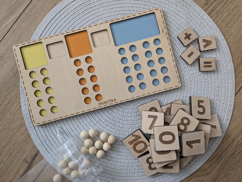 Pu&ograve; includere: Un tabellone di conteggio in legno con sezioni colorate e un'area blu per contare. Il tabellone include un set di tessere numeriche in legno, segni pi&ugrave;, meno, uguale e maggiore di. Ci sono anche palline di legno per contare.