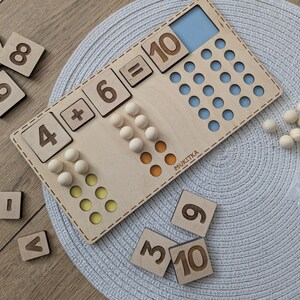 Pu&ograve; includere: Giocattolo di apprendimento in legno con un tabellone numerico e dei contatori. Il tabellone ha l'equazione 4 + 6 = 10 scritta su di esso. Ci sono anche tessere numeriche in legno con i numeri 1, 8, 9, 3, 10, 6 e 9.