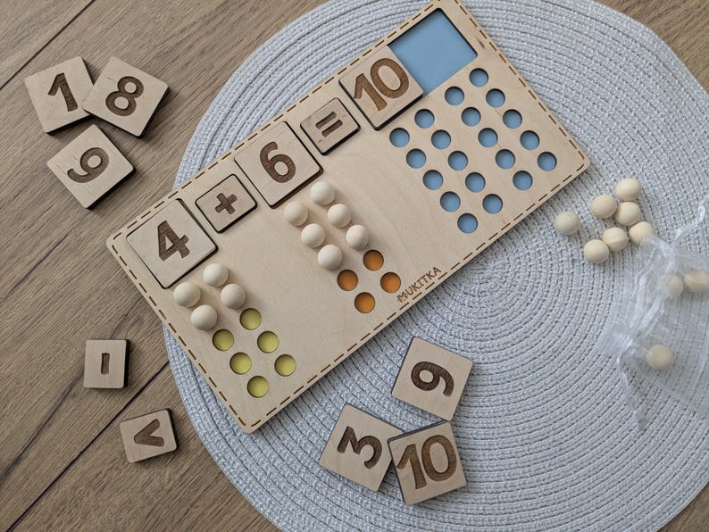 Pu&ograve; includere: Giocattolo di apprendimento in legno con un tabellone numerico e dei contatori. Il tabellone ha l'equazione 4 + 6 = 10 scritta su di esso. Ci sono anche tessere numeriche in legno con i numeri 1, 8, 9, 3, 10, 6 e 9.