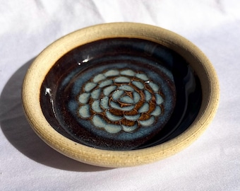 Puede incluir: Un plato redondo de cerámica con un borde beige claro texturizado y un esmalte interior marrón oscuro brillante. El centro presenta un patrón azul en forma de flor con pétalos superpuestos. El texto "Blue Rutile" aparece abajo a la derecha.