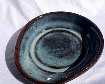 May include: A small, round, handmade ceramic dish with a dark brown rim and a glossy interior glaze featuring swirling patterns of blue, teal, and cream. The text 'Blue Rutile' is printed in the bottom right corner of the image.