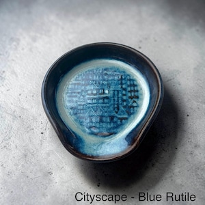 May include: A blue ceramic spoon rest with a cityscape design. The spoon rest is oval-shaped with a dark blue rim and a lighter blue interior. The text "Cityscape - Blue Rutile" is at the bottom.