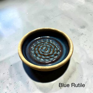 May include: A round ceramic dish with a matte beige exterior rim and a glossy interior featuring a deep blue glaze with a brown, flower-like embossed pattern in the center. The text Blue Rutile is printed in the bottom right corner of the image.