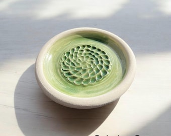 Pode incluir: Um prato redondo de cerâmica verde celadon com um design texturizado em forma de flor no centro. O prato tem uma borda branca e o texto "Celadon Green" é visível na imagem.