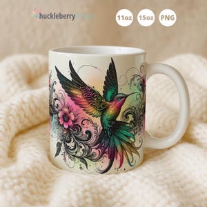 Op de afbeelding: Witte keramische mok met een kleurrijk ontwerp van een kolibrie en bloemmotieven. De mok heeft een gebogen handvat en de tekst "huckleberry dreams" is zichtbaar. Verkrijgbaar in 312 ml en 425 ml maten.