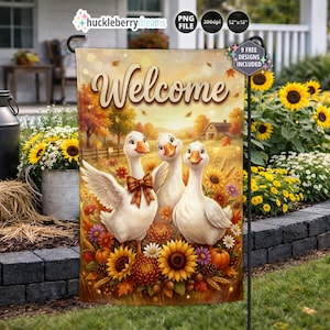 Puede incluir: Una bandera de jardín rectangular con la palabra Welcome en letra cursiva sobre tres gansos blancos rodeados de flores otoñales, incluyendo girasoles, crisantemos naranjas y calabazas, con un paisaje de atardecer de fondo.